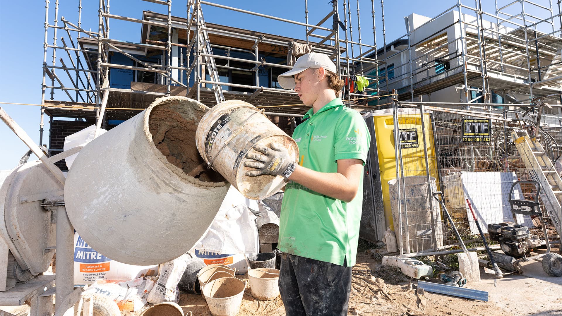 A construction worker in a green shirt and cap pours a bucket of material into a concrete mixer on a busy building site, with scaffolding and various construction materials in the background.