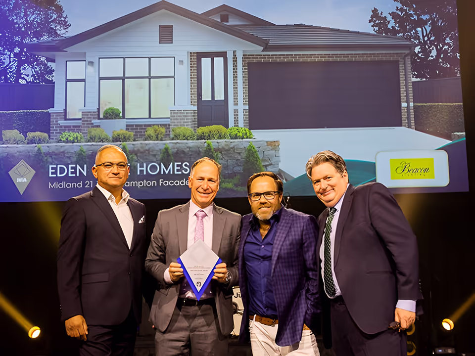 Four men in suits stand on stage smiling, one holding an award. Behind them, a screen displays a house image and the text Eden Brae Homes and Midland 21 Hampton Facade. Stage lighting highlights the group.
