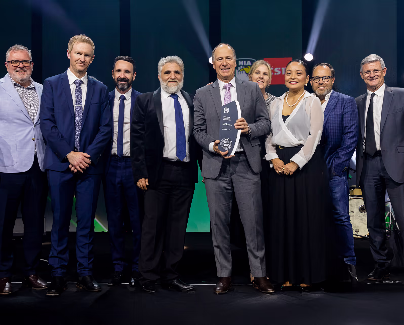 A group of ten people in formal attire stand on stage, smiling. The person in the center holds a trophy or award. There are stage lights and colorful backgrounds behind them, suggesting an awards ceremony or celebration.