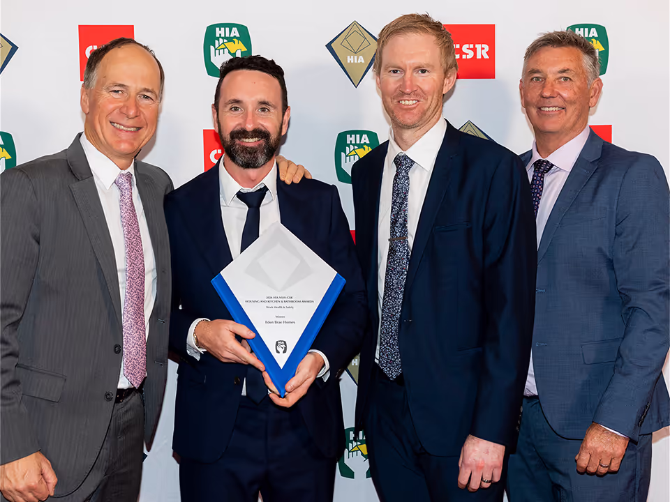 Four men in suits pose together at an awards event; one man in the middle holds a framed certificate. The background displays various sponsor logos.