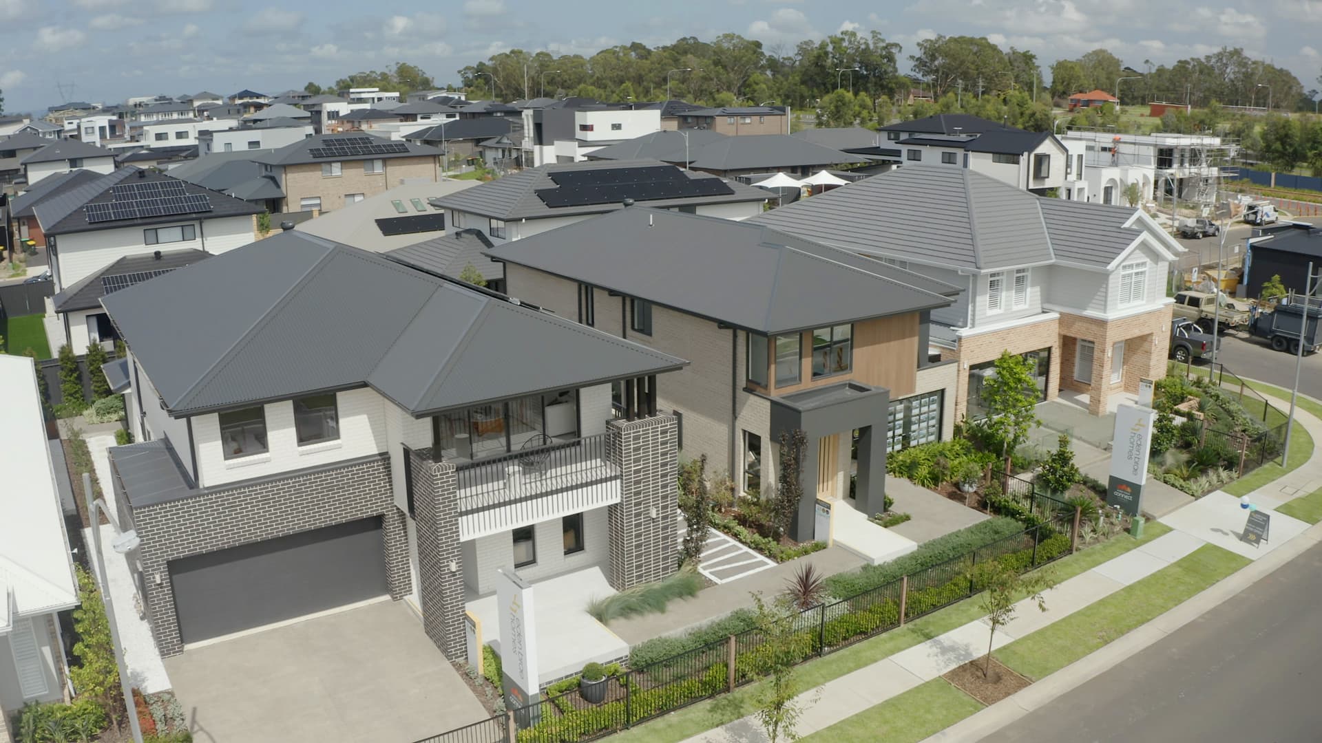 Aerial view of a modern suburban neighborhood with large, two-story houses, neatly landscaped yards, and solar panels on some rooftops under a partly cloudy sky.