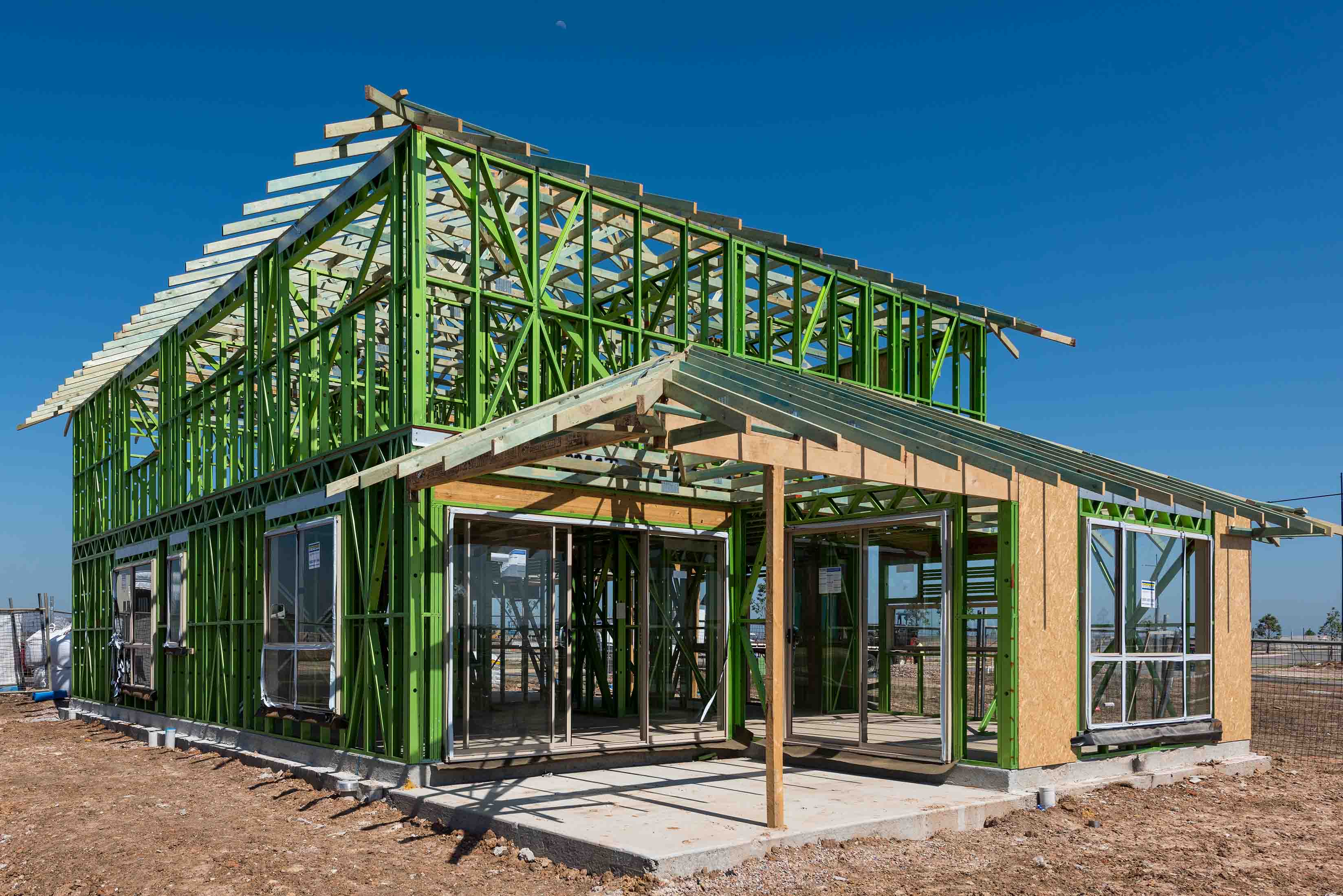 A house under construction with a green steel frame structure, partially covered roof, and installed windows, set on a bare plot of land under a clear blue sky.