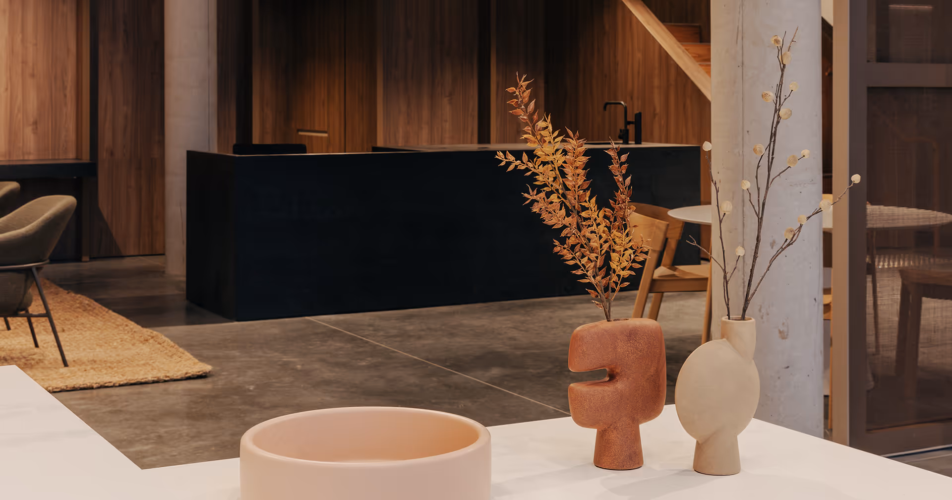 Modern interior with minimalist decor, featuring two sculptural vases with dried branches on a white surface, a round shallow bowl, and a sleek, dark wood kitchen in the background. Warm, earthy tones throughout.