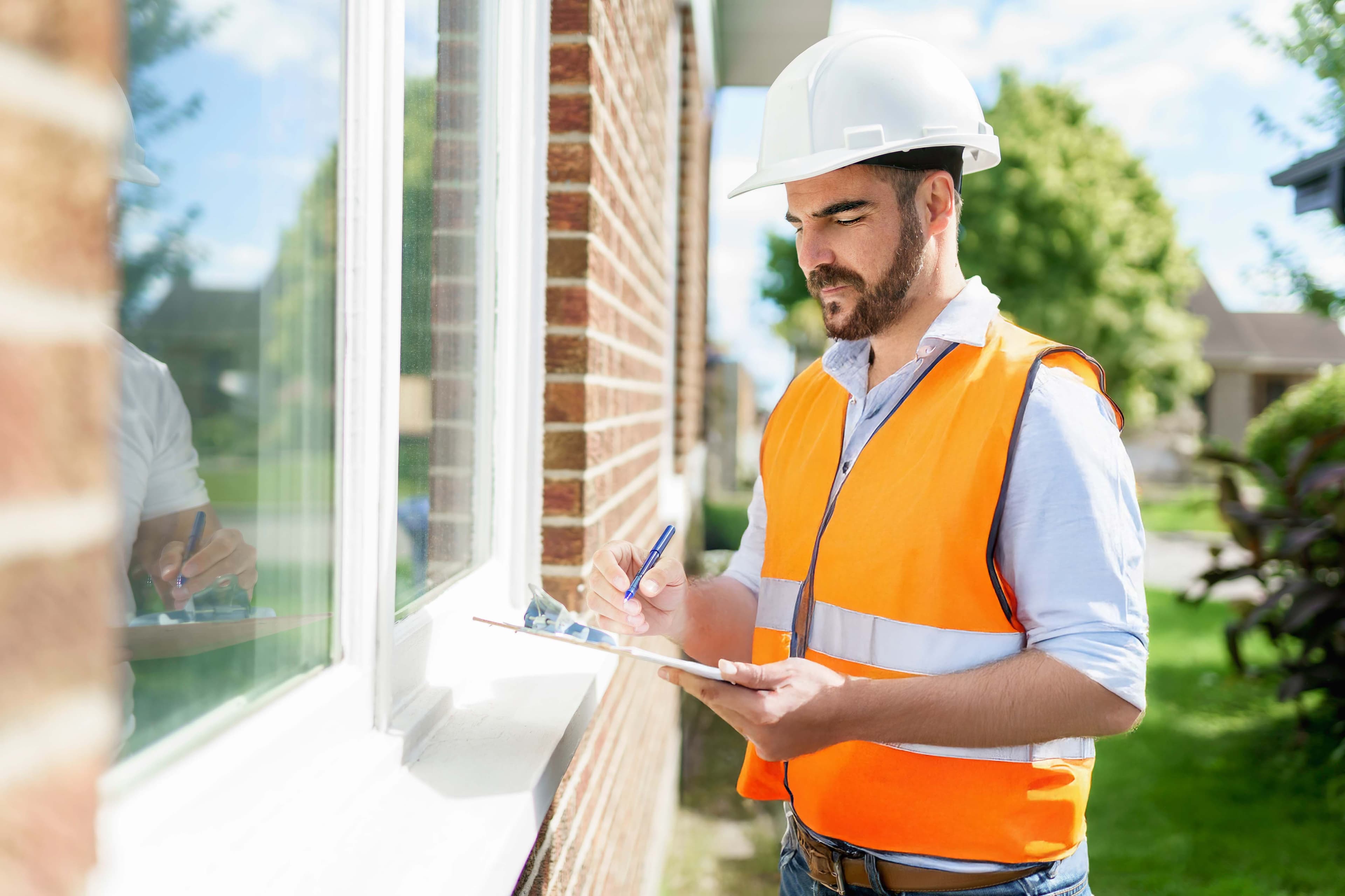 A man wearing a white safety helmet and orange reflective vest stands outside a brick building, writing on a clipboard while inspecting a window. Trees and other houses are visible in the background.