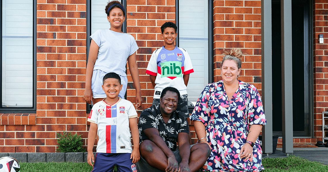 Five people, including two adults and three children, smile and pose together outside a brick house. Some children wear rugby jerseys and a rugby ball is visible on the grass in the foreground.