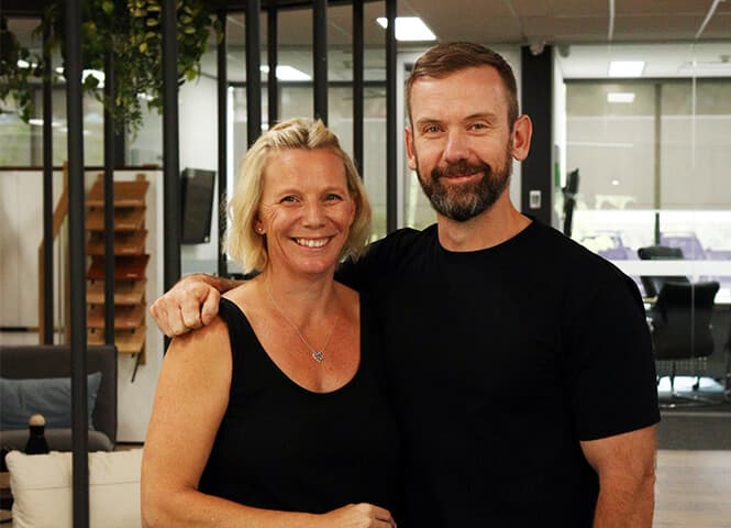 A woman and a man stand indoors in an office setting, smiling at the camera. The man has his arm around the womans shoulders. Both are casually dressed in black shirts. The background features desks and windows.