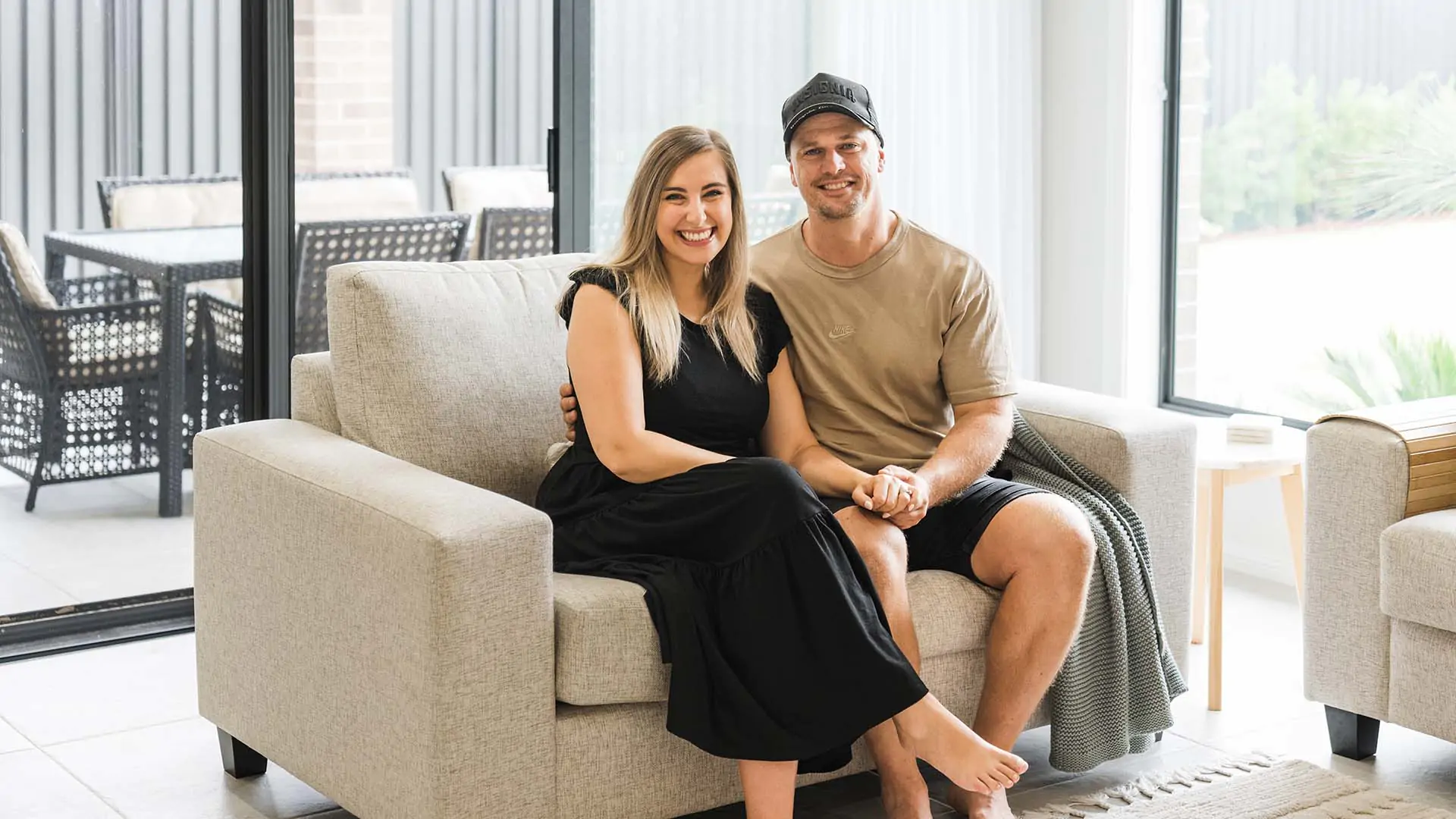 A smiling couple sits closely together on a beige sofa in a bright, modern living room. The woman wears a black dress, and the man wears a tan shirt, black shorts, and a cap. Large windows provide natural light.